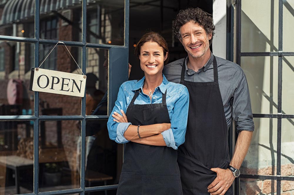Two People Stood Next to Each Other Smiling at The Door Entrance of an Open Shop